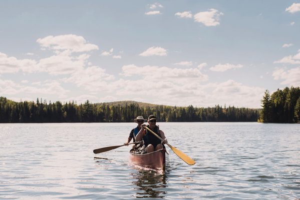 custom pak nz partners, Two people are canoeing on a calm lake surrounded by forest. The sky is bright with scattered clouds, and the water reflects the light softly